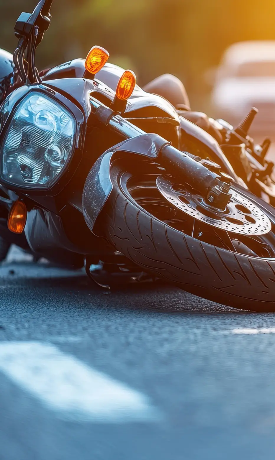 A fallen black motorcycle lies on its side on a sunlit road, with glowing orange indicators and blurred cars in the background, evoking tension and urgency.