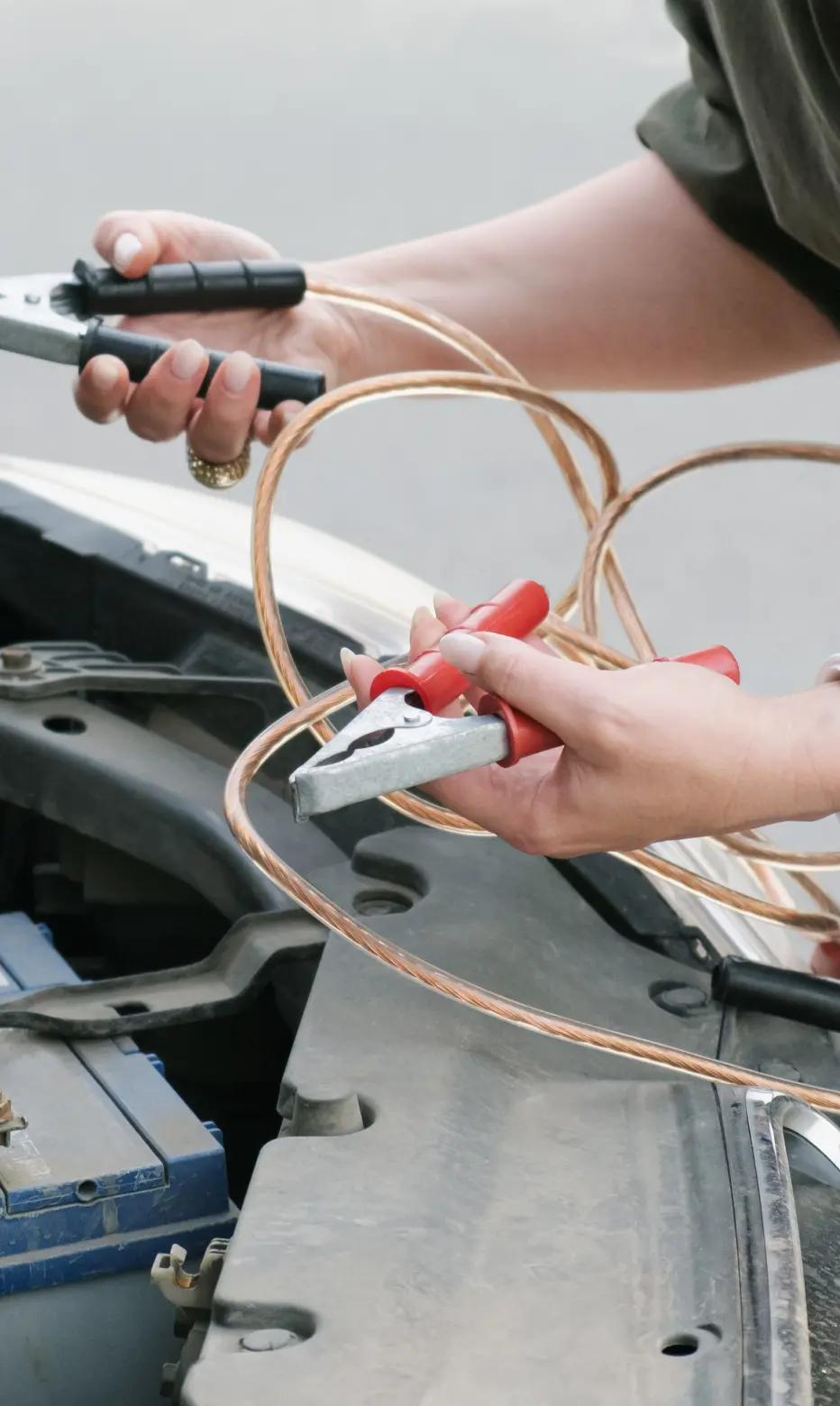 A person holds jumper cables above an open car hood, preparing to connect them to a battery. The scene conveys urgency and readiness for action.