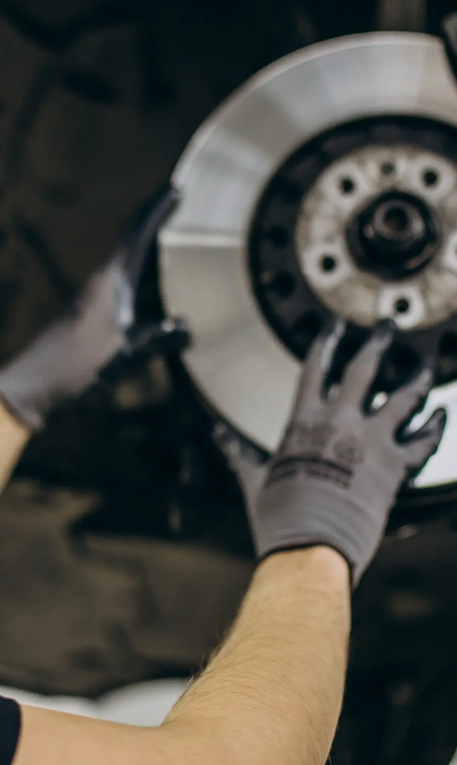 A person wearing gloves installs a shiny, new brake disc onto a vehicle. The setting is a workshop, conveying focus and precision in automotive repair.