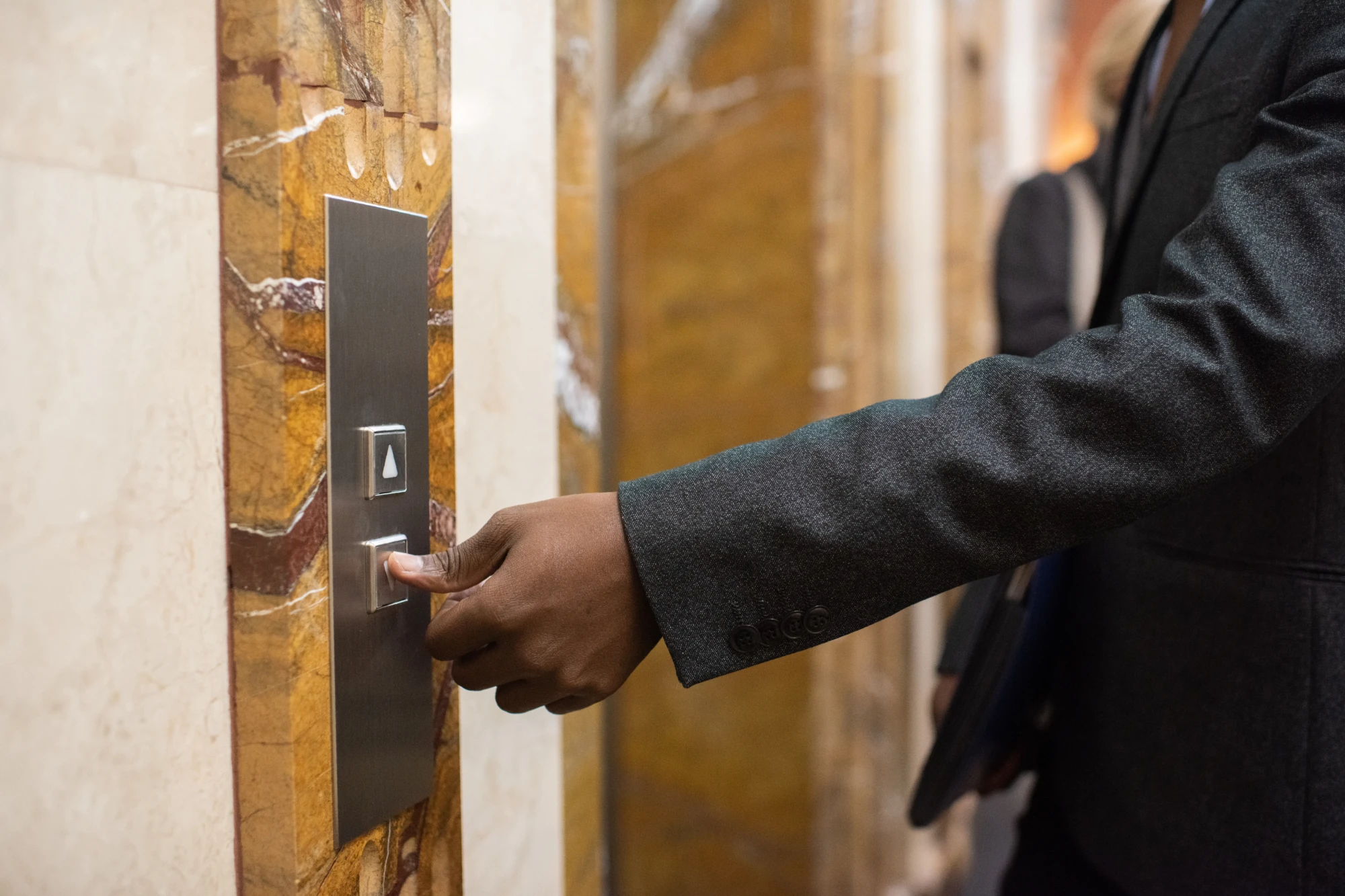 A person in a dark suit jacket presses an elevator button on a marble wall, conveying a sense of action and daily business routine.