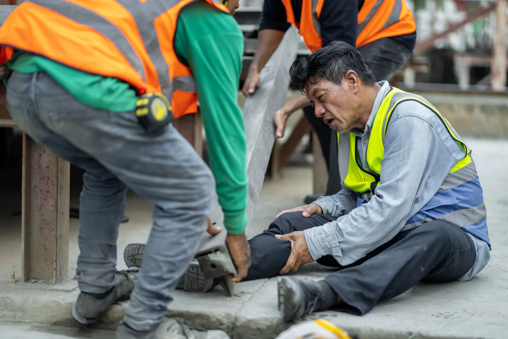 A construction worker in a high-visibility vest sits on the ground, holding his injured leg, surrounded by concerned colleagues in safety gear. The scene conveys urgency.