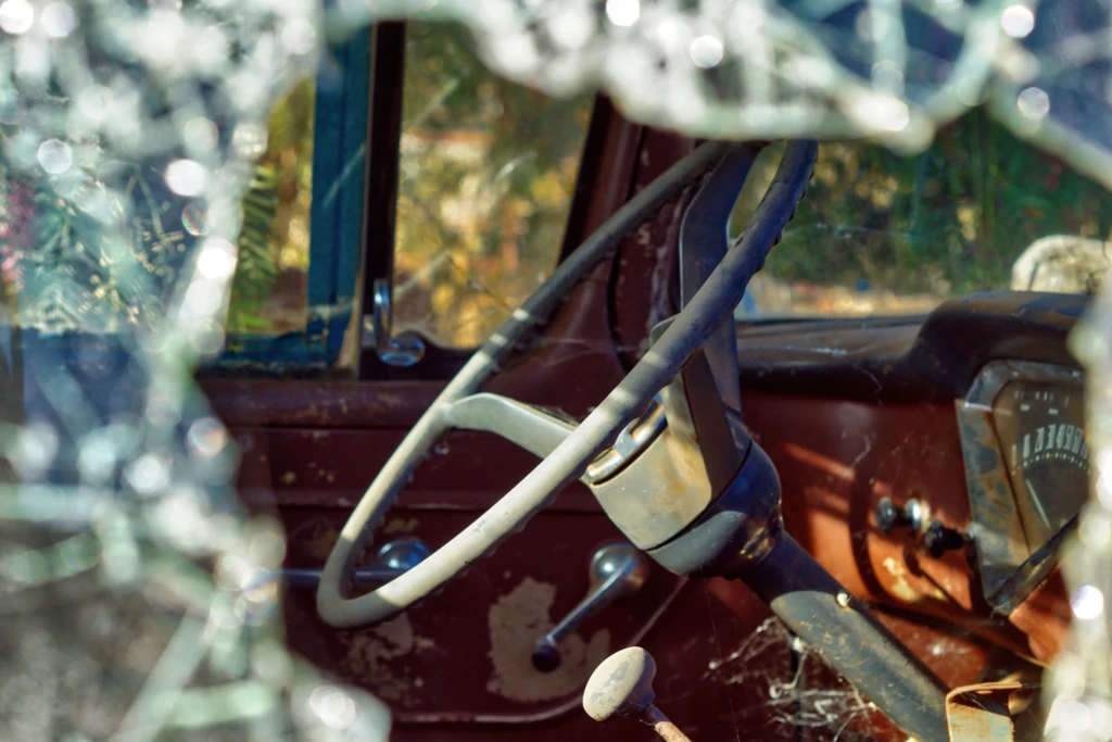 Close-up of a weathered car interior seen through a shattered window. The scene is filled with nostalgia, highlighting a rusty steering wheel and dashboard.