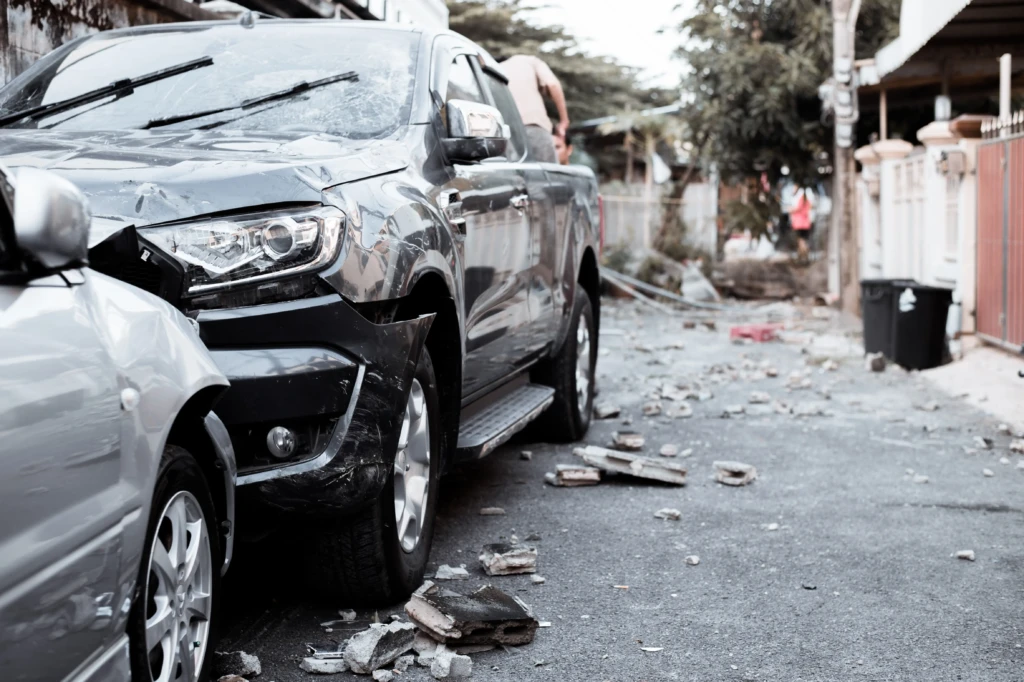 A man sits on the ground in front of a badly damaged car, his head resting on his arm, conveying distress and exhaustion in a sunny outdoor setting.
