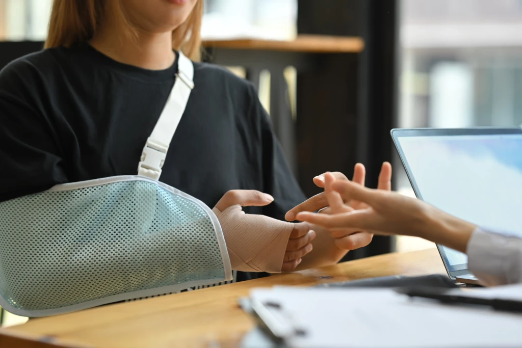 A person wearing a black shirt with an arm in a sling is having their bandaged hand examined by another individual across a table, conveying a clinical setting.