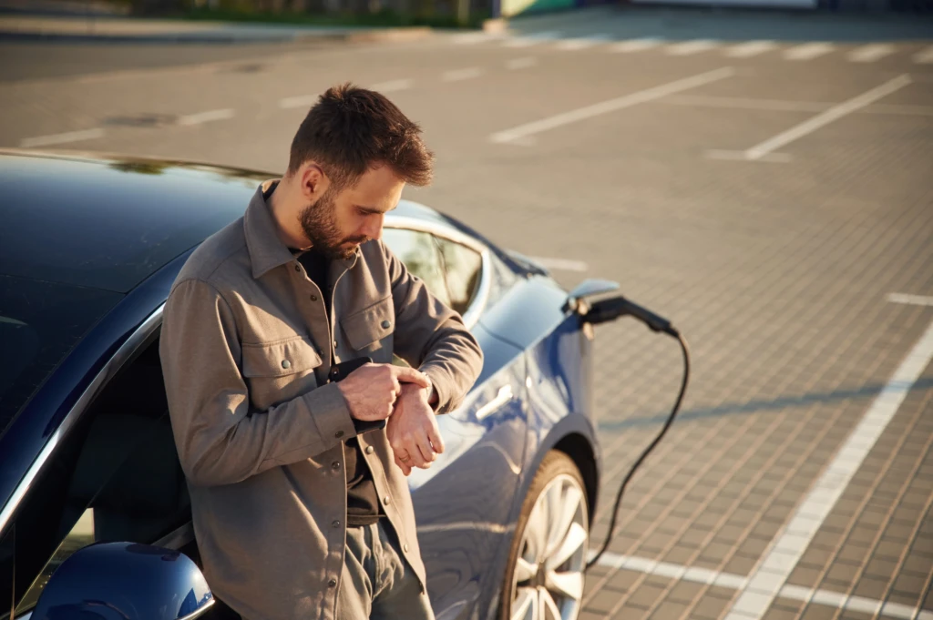 A man stands by a blue electric car plugged into a charging station, looking at his watch in a sunlit parking lot, conveying patience and anticipation.