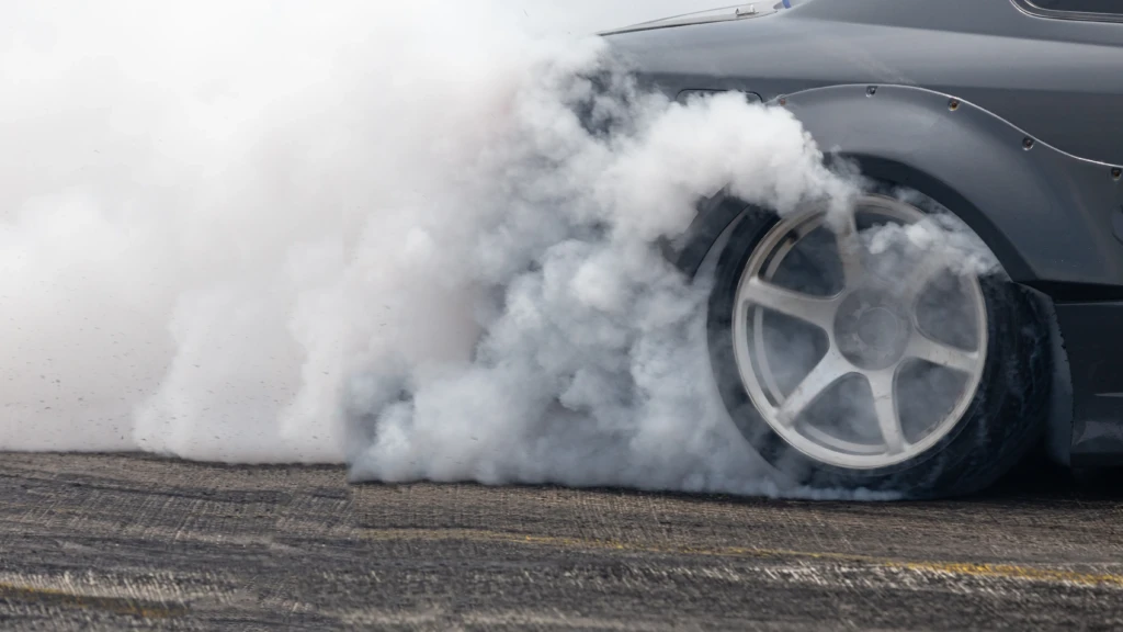 Close-up of a car tire spinning on a track, creating thick white smoke from the friction. The image conveys speed, power, and the thrill of drifting.