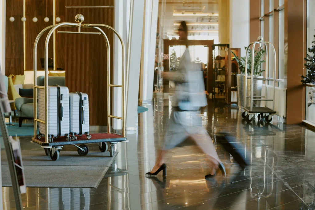 A person in motion pulls a suitcase across a glossy hotel lobby. Two luggage carts with suitcases are nearby, adding a sense of busy travel.