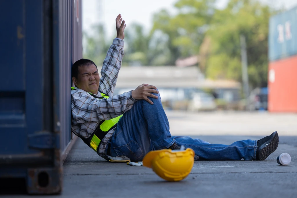 A worker in a plaid shirt and safety vest sits on the ground, grimacing in pain. A yellow hard hat and cup lie nearby. The scene appears tense.