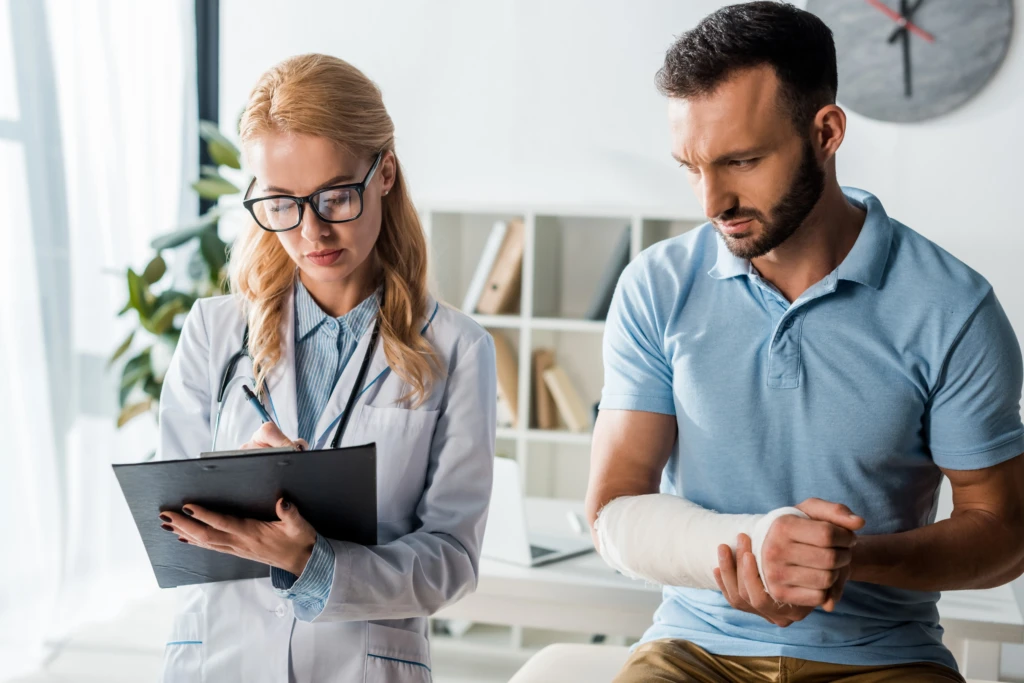A doctor in a white coat and glasses writes on a clipboard while a concerned man in a blue shirt, with a bandaged arm, looks on in a medical office.