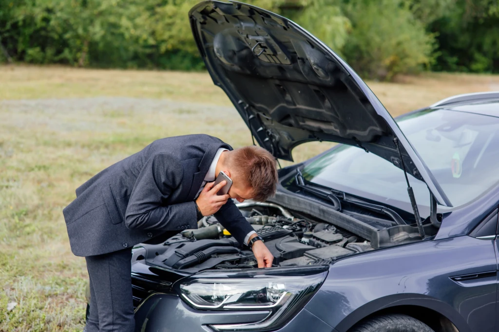 A man in a suit examines a car engine under an open hood while talking on the phone, looking concerned. The car is parked in a grassy area.