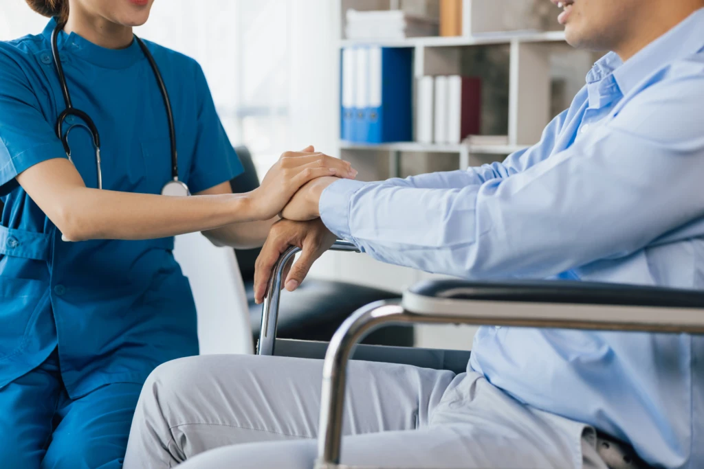 A nurse in blue scrubs empathetically holds the hand of a person sitting in a wheelchair, conveying support and care in a clinical setting.