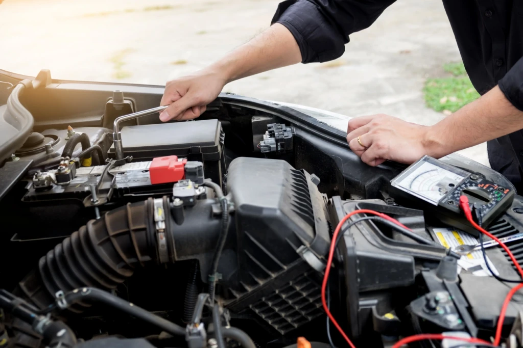 A person works on a car engine, using a wrench and a multimeter with red cables attached. The scene conveys focus and technical expertise.