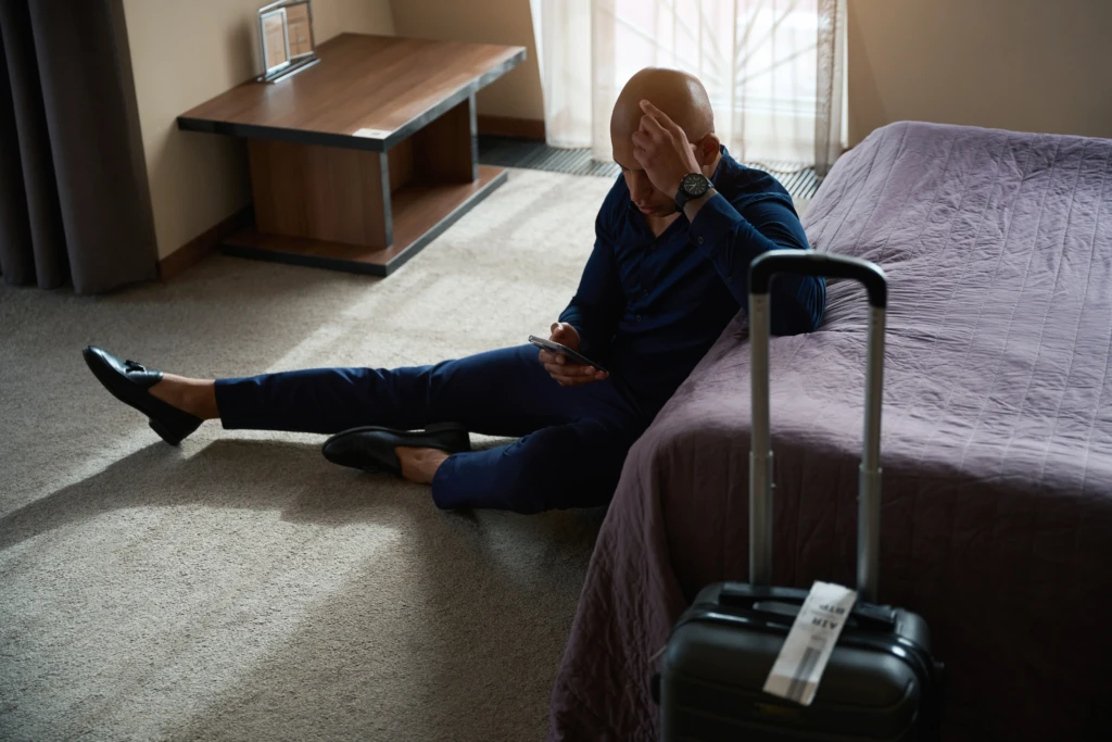 Man in a blue suit sitting on a hotel room floor, looking at his phone with a frustrated expression. A suitcase stands beside him near the bed.