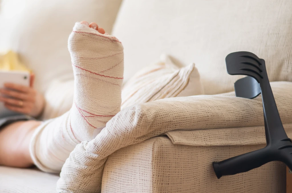 A person with a bandaged leg rests it on a cushion on a beige couch, holding a smartphone. A black crutch leans nearby, suggesting recovery.
