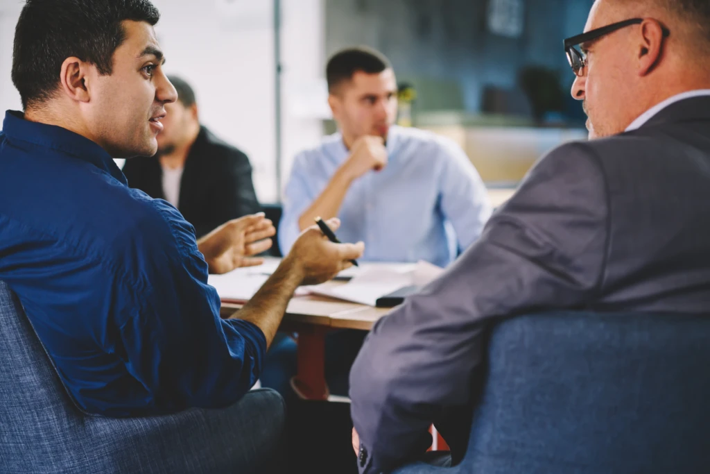 Four men engaged in a serious discussion around a table, one gesturing with a pen. The setting is a modern office, conveying a collaborative tone.