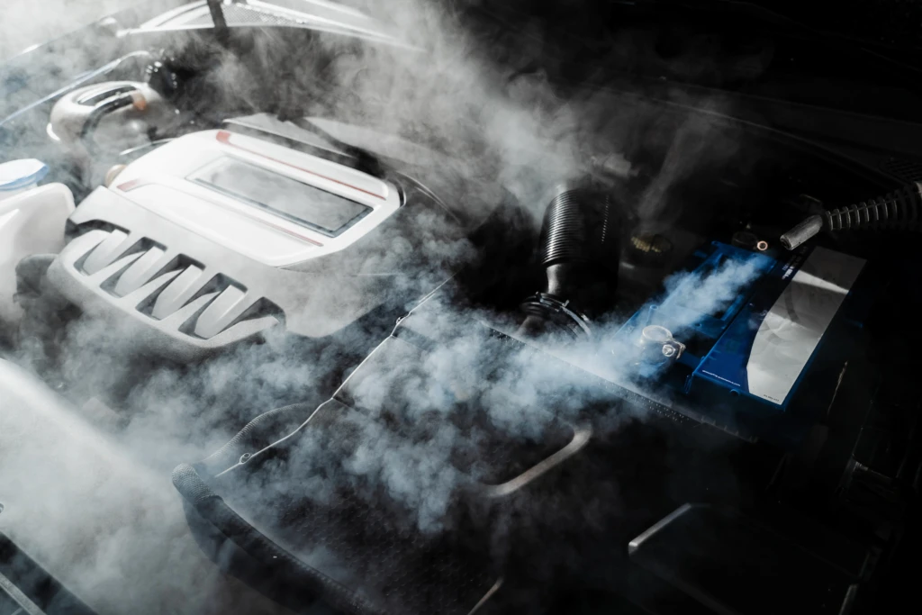 Car engine emitting smoke from under the hood, creating a dramatic, tense atmosphere. Visible engine parts contrast against the white, swirling smoke.