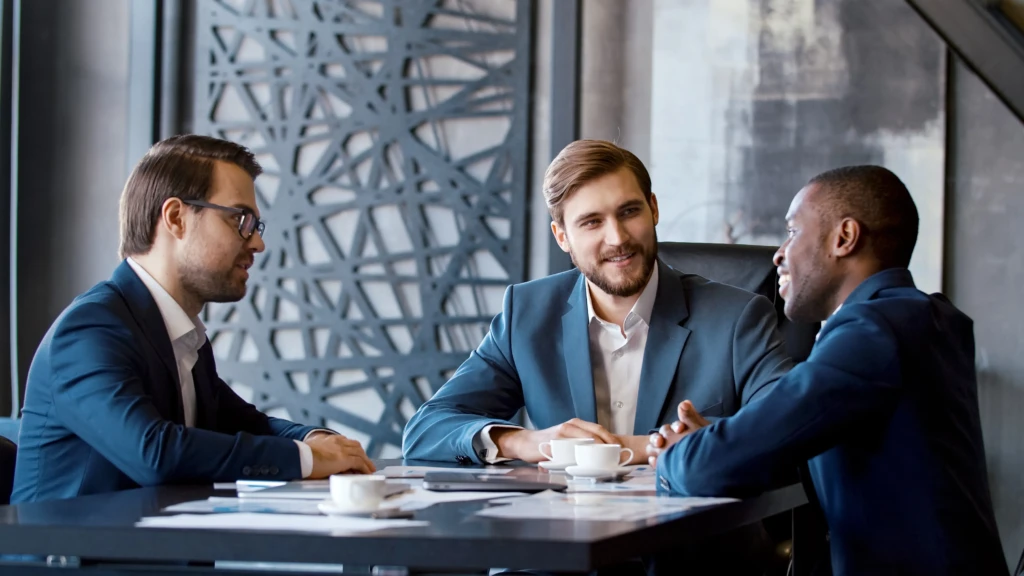 Three men in suits sit around a table in a modern office, engaged in a friendly conversation. Documents and coffee cups are on the table.